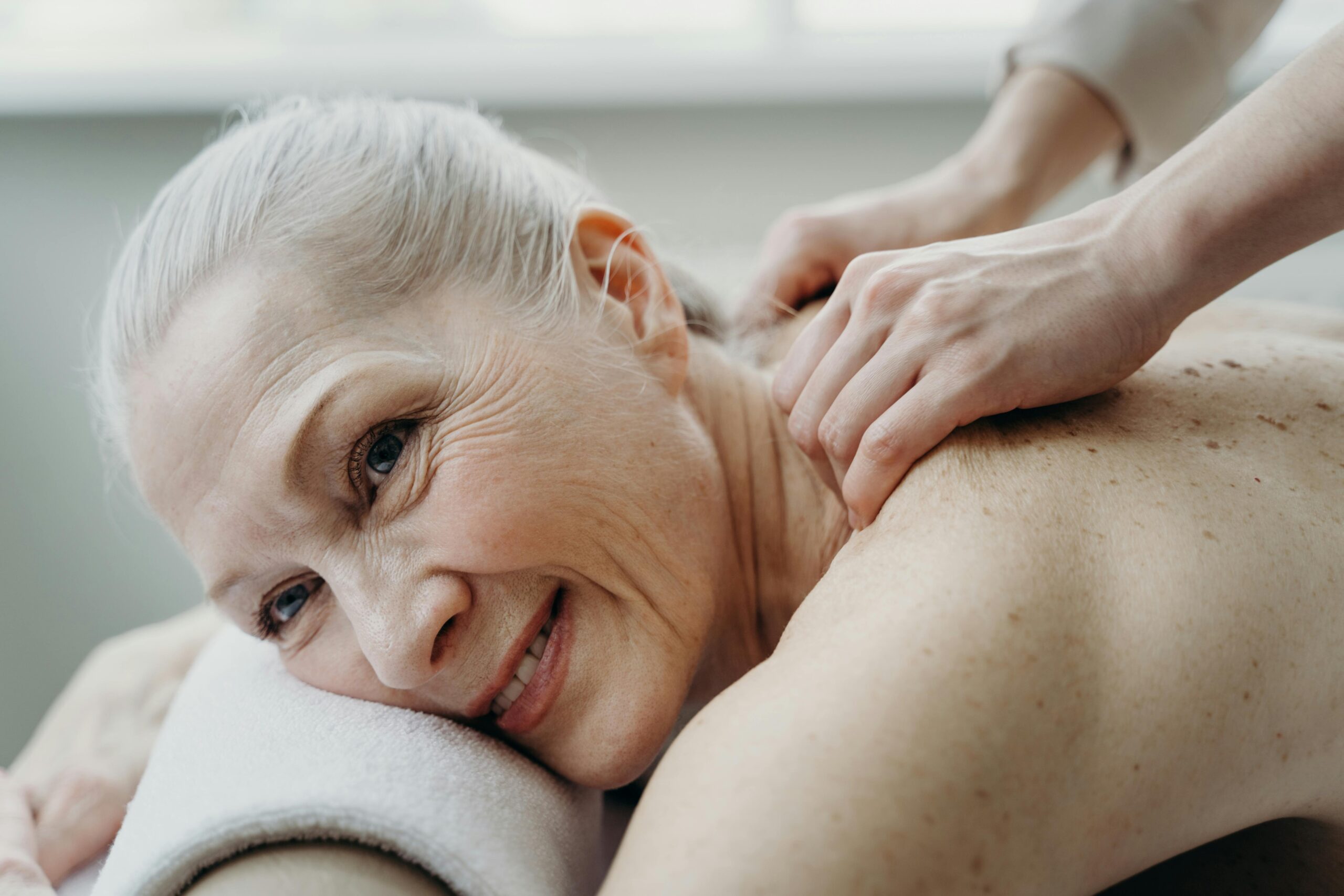 A senior woman with white hair smiles while receiving a therapeutic back massage indoors.